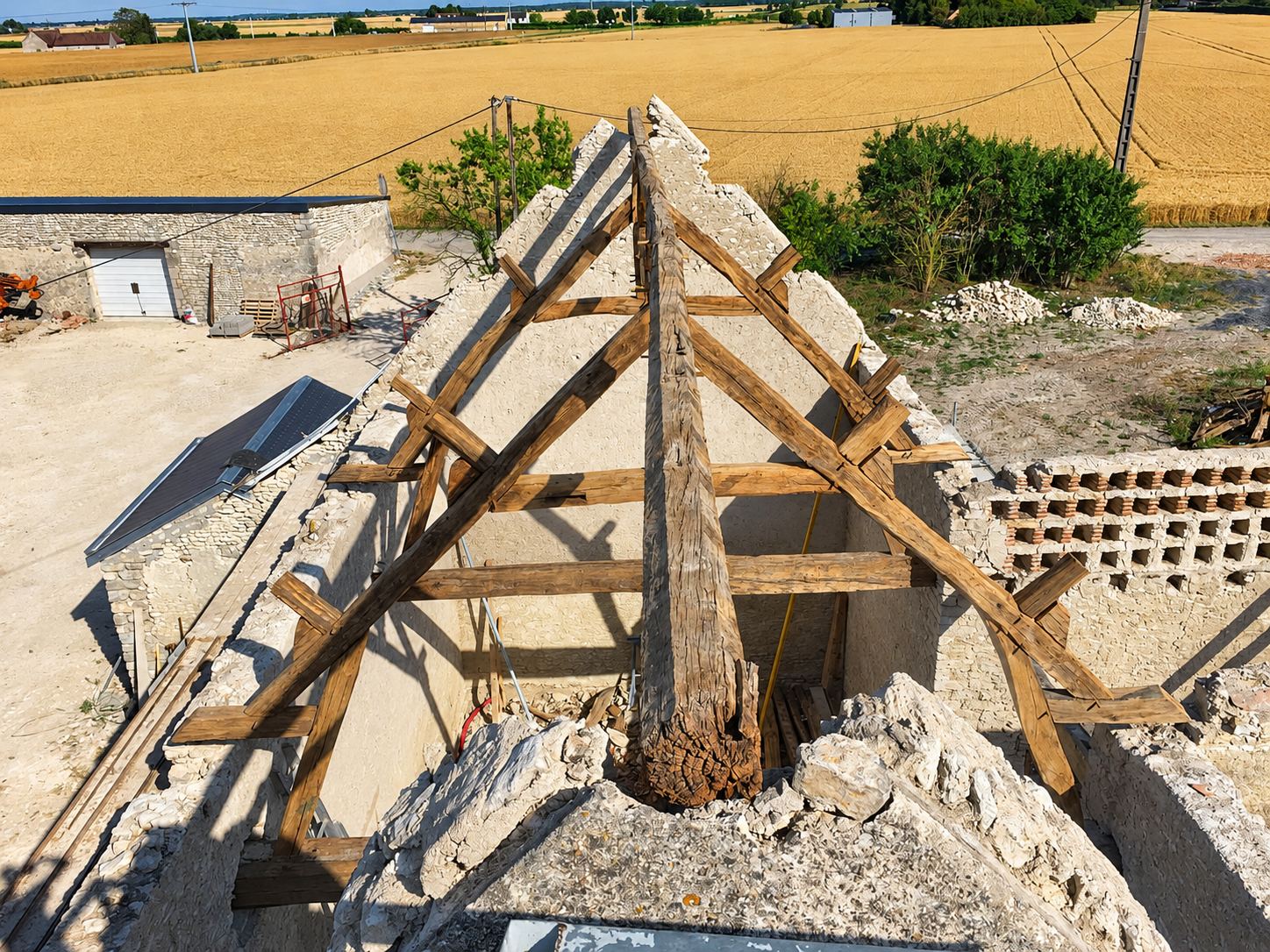 Vue depuis le haut d'une vieille charpente en chêne mise à nu sur les murs en pierre d'une grange en rénovation, au milieu de champs de blé.