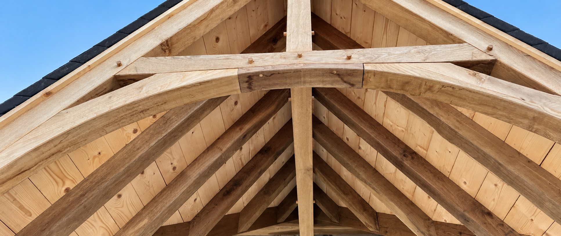 Vue en contre-plongée d'une charpente traditionnelle en chêne avec poutres cintrées, chevilles en bois et voligeage clair sous un ciel bleu.