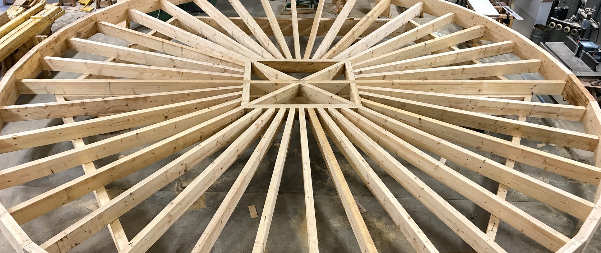 Structure de charpente circulaire en bois clair avec chevrons rayonnants, assemblée au sol dans un atelier de menuiserie.