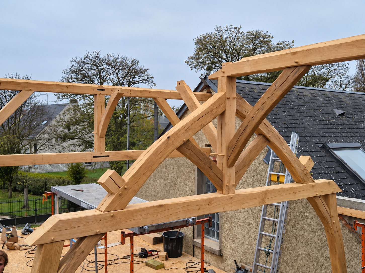 Une charpente en chêne massif aux assemblages apparents est en cours de montage au sommet d'une maison, avec une échelle et du matériel de chantier autour.