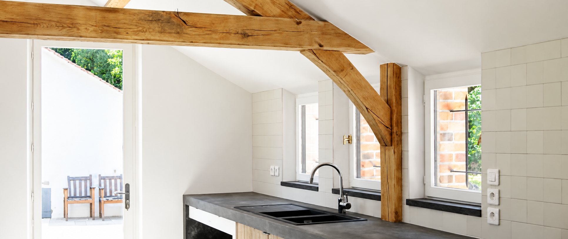 Cuisine lumineuse aux murs blancs et carrelage clair, avec poutres en chêne apparentes, plan de travail en béton gris et évier noir.