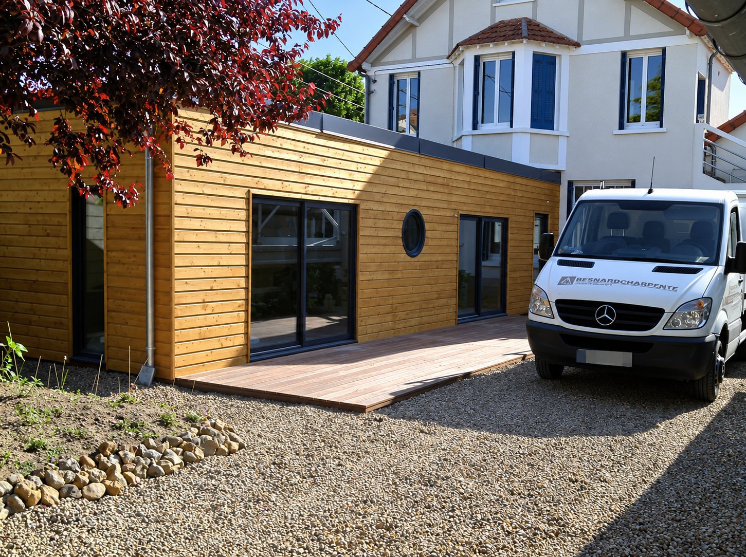 Une extension en bardage bois avec grandes baies vitrées et terrasse en bois, devant une maison, avec une camionnette blanche d'entreprise de charpente garée sur le gravier.