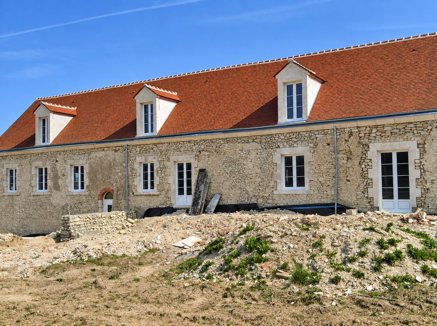 Ancien bâtiment en pierre en cours de rénovation, avec une toiture neuve en tuiles rouges, des lucarnes et un terrain de chantier au premier plan.