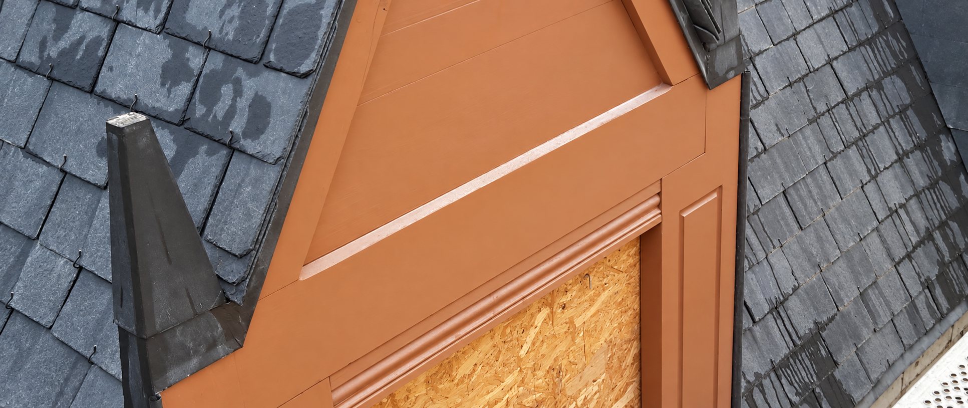 Gros plan d'une lucarne peinte en brun, dont la fenêtre est provisoirement fermée par un panneau de bois OSB, entourée d'un toit en bardeaux gris foncé.