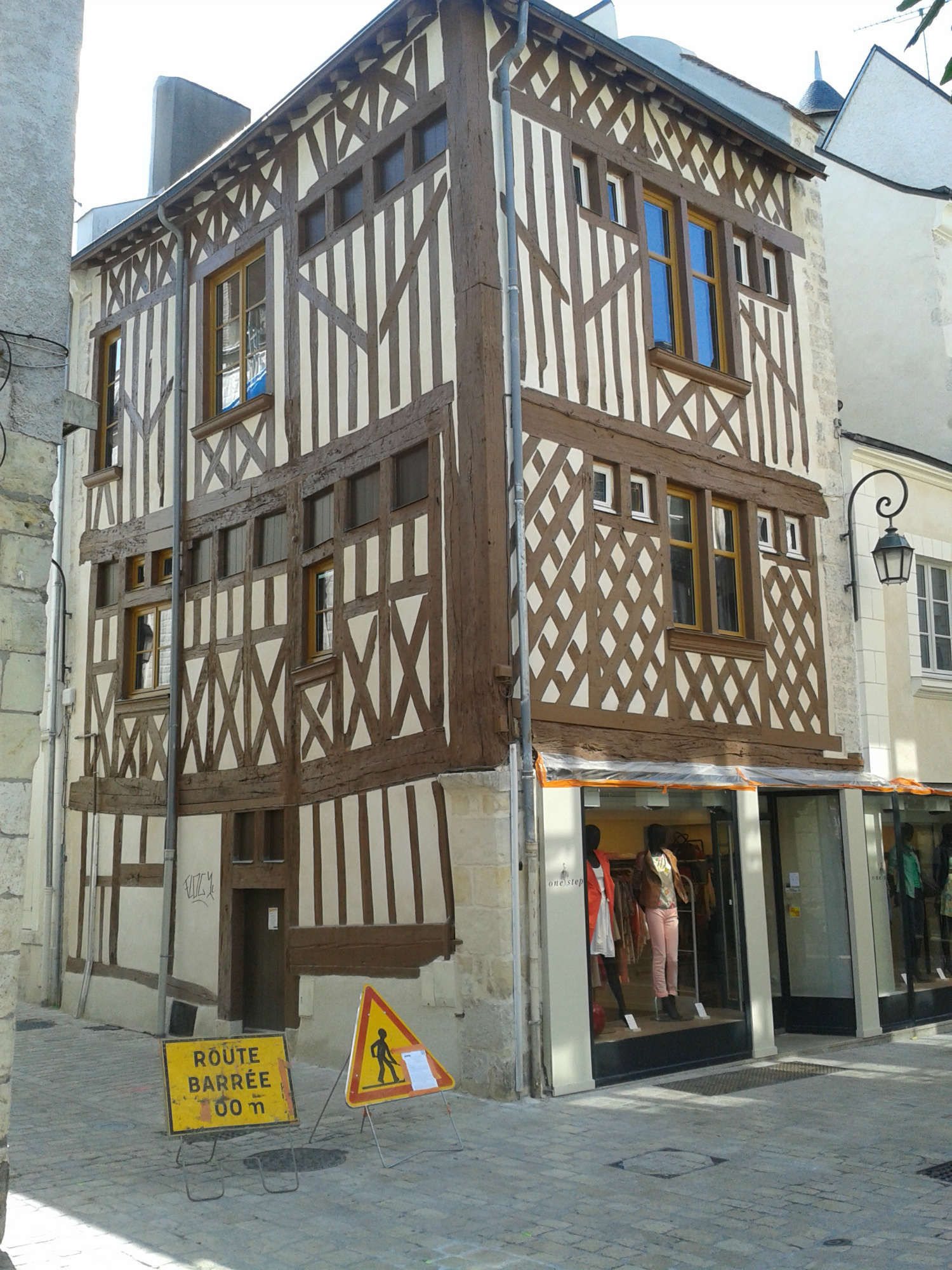 Façade d'une maison à colombages de trois étages dont le rez-de-chaussée est une boutique de vêtements, avec un panneau de route barrée devant.
