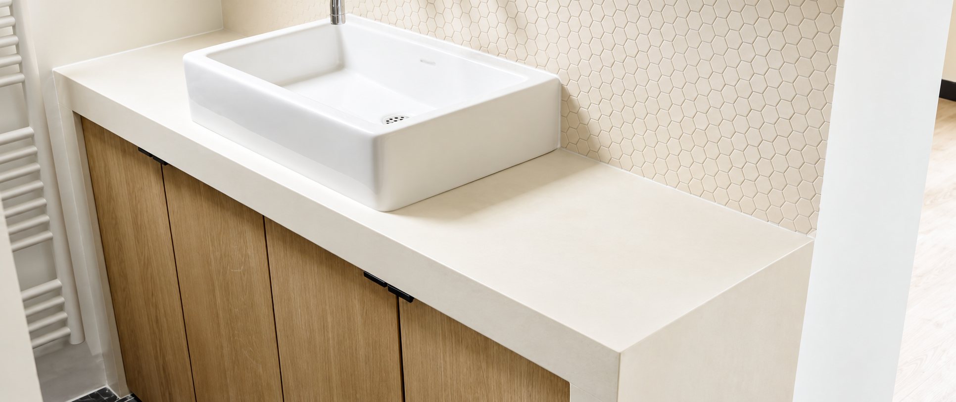 Vasque blanche rectangulaire posée sur un plan de travail clair en béton ciré, au-dessus d'un meuble en bois et devant un mur en mosaïque hexagonale.