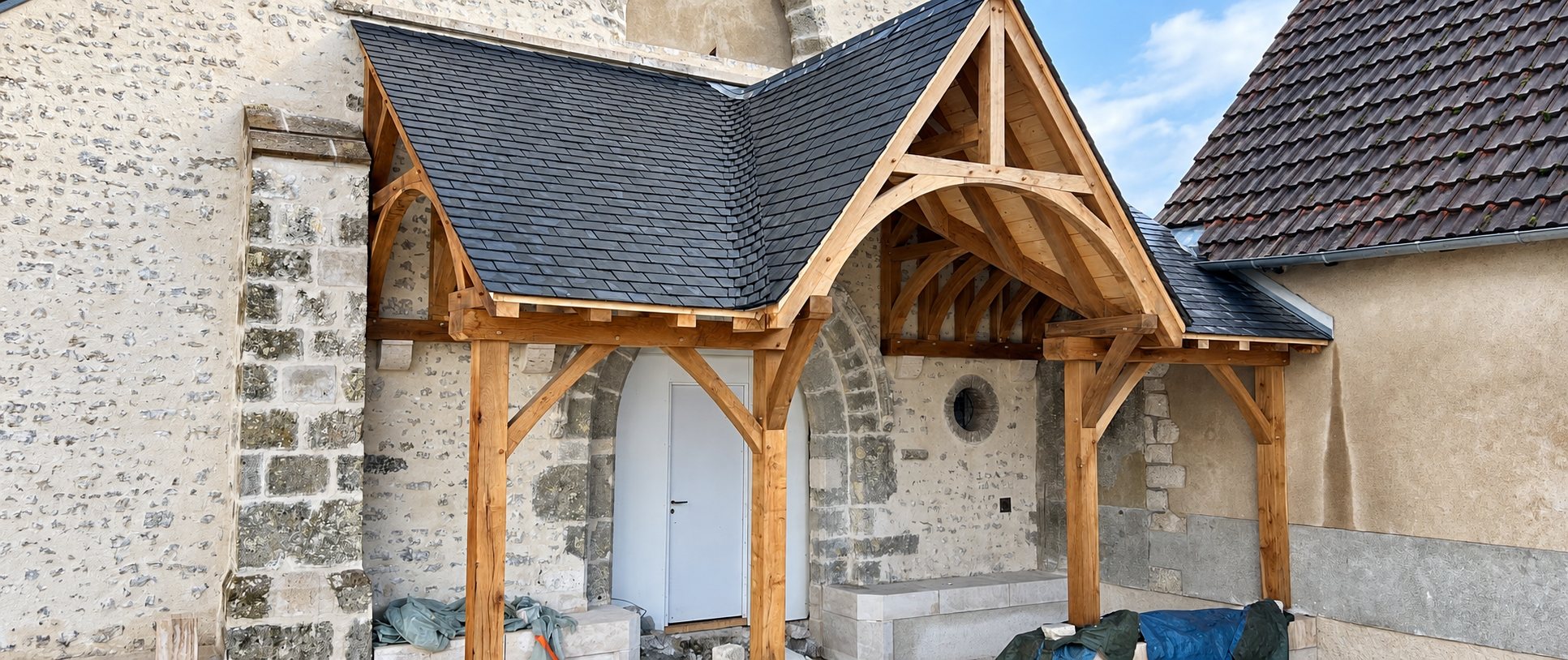 Un porche neuf en charpente de chêne apparente avec toit en ardoise protège une porte blanche sur la façade en pierre d'une église.