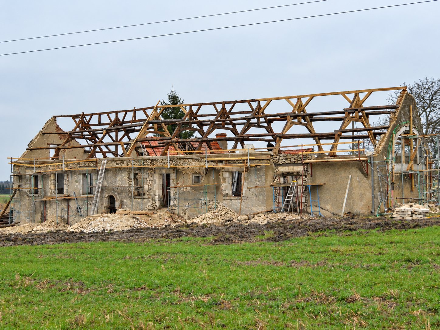 Chantier de rénovation d'un long bâtiment en pierre dont la charpente en bois est mise à nu, entouré d'échafaudages et de gravats, au milieu d'un champ.